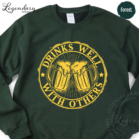 A green sweatshirt with a yellow graphic design on it. The design features two beer mugs with a text that says "DRINKS WELL WITH OTHERS".