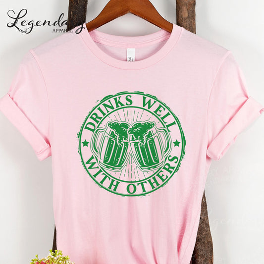 A pink t-shirt with a green graphic design that says "DRINKS WELCOME WITH OTHERS" on it.
