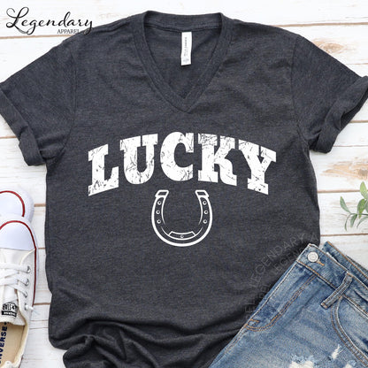 A gray t-shirt with the word "LUCKY" printed on it, along with a horseshoe graphic, is displayed on a wooden surface.
