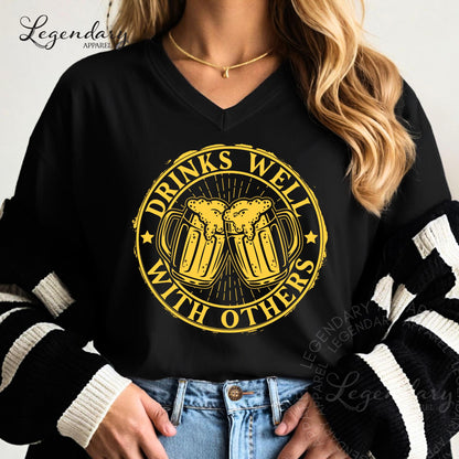 A woman wearing a black shirt with a graphic design that says "DRINKS WELL WITH OTHERS" in yellow and black.