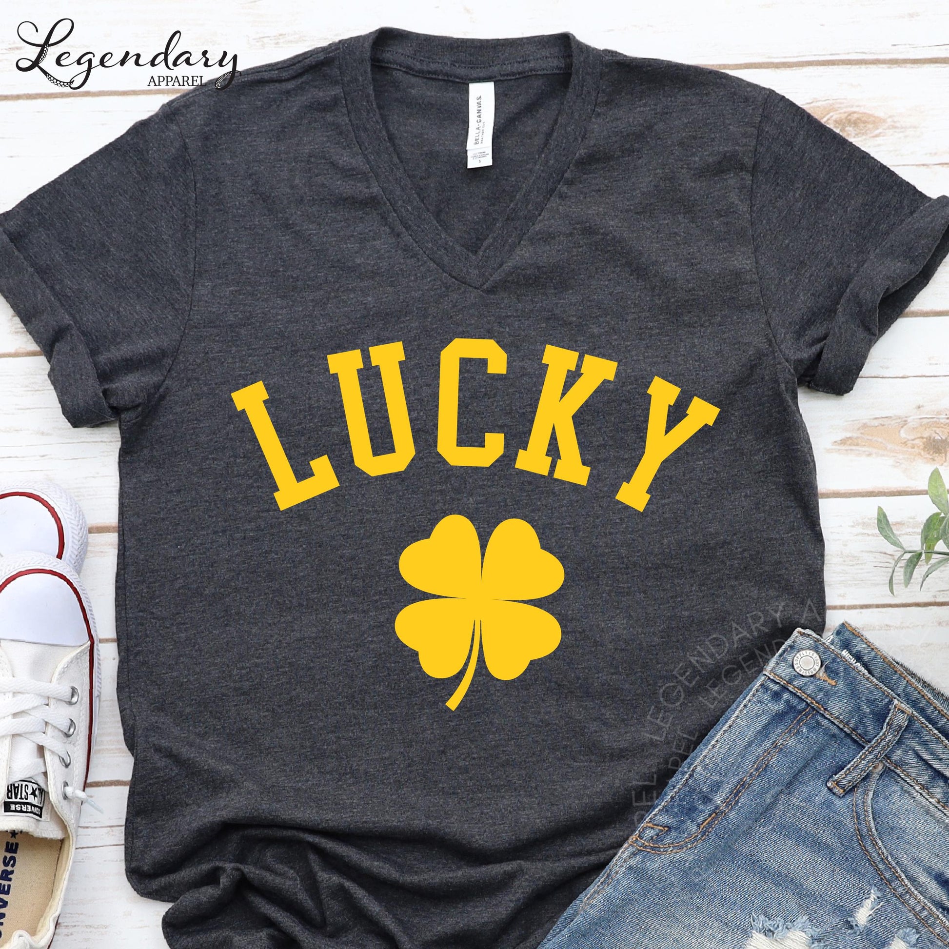 A gray t-shirt with the word "Lucky" printed in yellow letters, along with a yellow clover symbol, is displayed on a wooden surface.