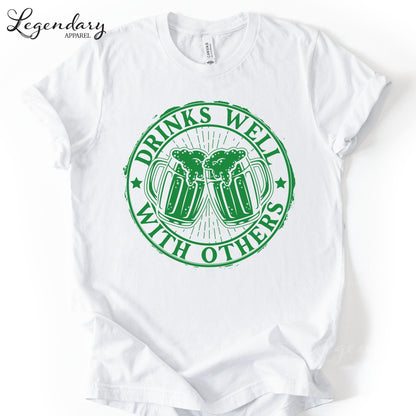 A white t-shirt with a green circular graphic design that says "DRINKS WELL WITH OTHERS" and features two beer mugs with foam on top.