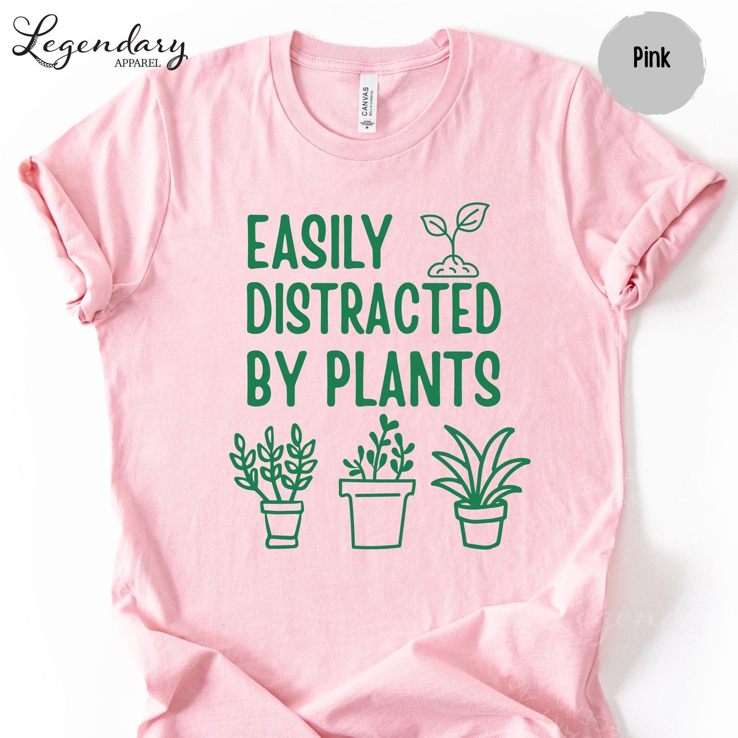 A pink t-shirt with a graphic design featuring a plant and the text "EASILY DISTRACTED BY PLANTS" printed on it.
