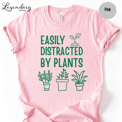 A pink t-shirt with a graphic design featuring a plant and the text "EASILY DISTRACTED BY PLANTS" printed on it.
