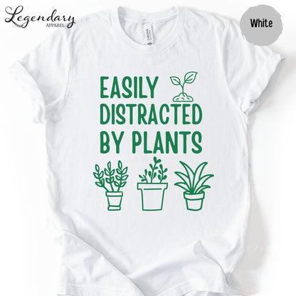 A white t-shirt with a graphic design featuring a green plant and the text "EASILY DISTRACTED BY PLANTS" printed on it.