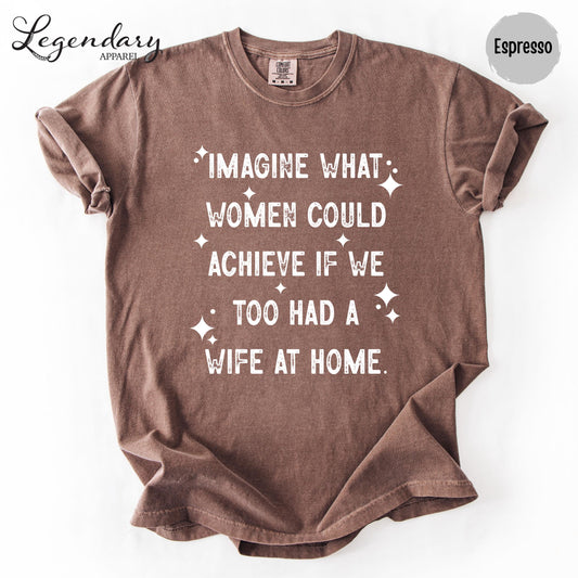 a brown t-shirt with white text printed on it. The text reads "IMAGINE WHAT WOMEN COULD ACHIEVE IF WE TOO HAD A WIFE AT HOME."