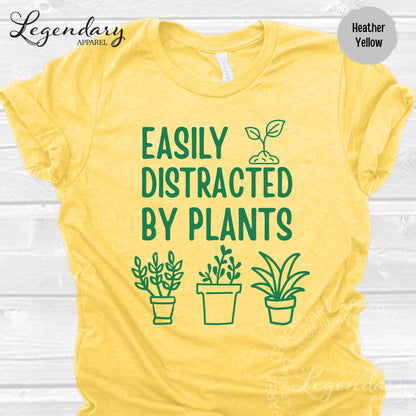 The image is a yellow t-shirt with a graphic design featuring a plant and the text "EASILY DISTRACTED BY PLANTS" printed on it.