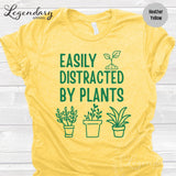The image is a yellow t-shirt with a graphic design featuring a plant and the text "EASILY DISTRACTED BY PLANTS" printed on it.
