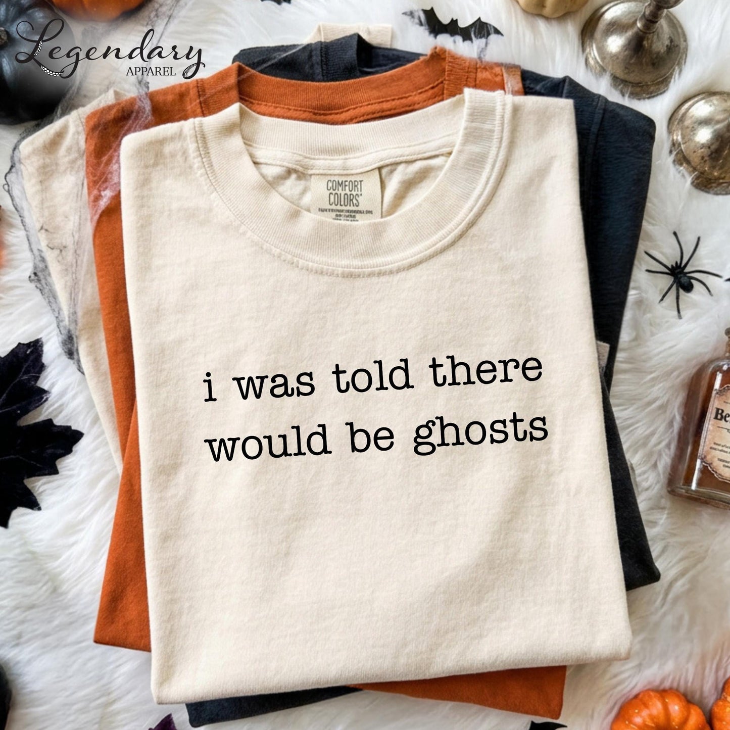 a white t-shirt with the text "I was told there would be ghosts" printed on it, along with a label that reads "Comfort Colors". The t-shirt is displayed on a white surface, surrounded by various Halloween-themed items such as a bottle, a pumpkin, and a spider.