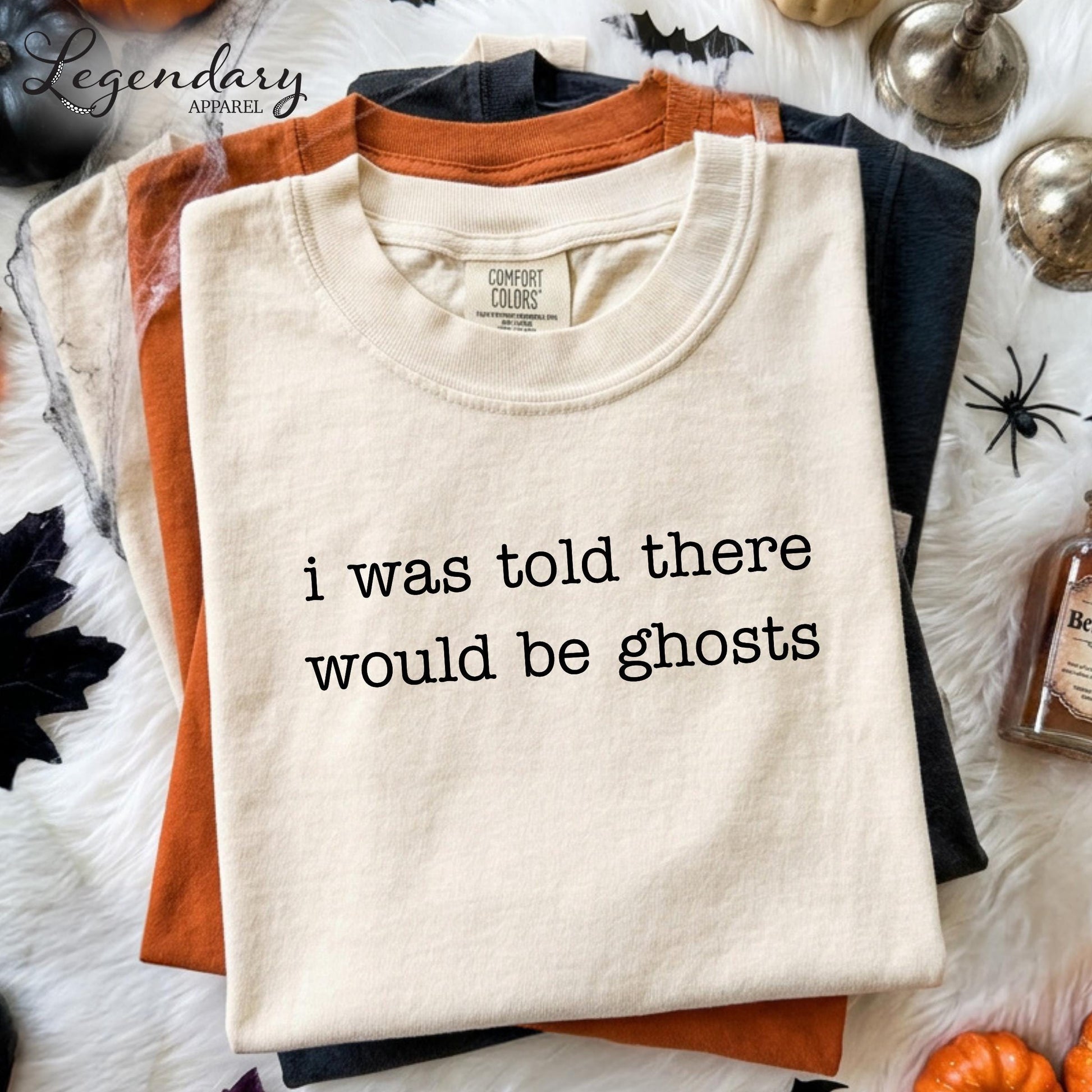 a white t-shirt with the text "I was told there would be ghosts" printed on it, along with a label that reads "Comfort Colors". The t-shirt is displayed on a white surface, surrounded by various Halloween-themed items such as a bottle, a pumpkin, and a spider.