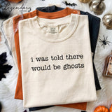 a white t-shirt with the text "I was told there would be ghosts" printed on it, along with a label that reads "Comfort Colors". The t-shirt is displayed on a white surface, surrounded by various Halloween-themed items such as a bottle, a pumpkin, and a spider.