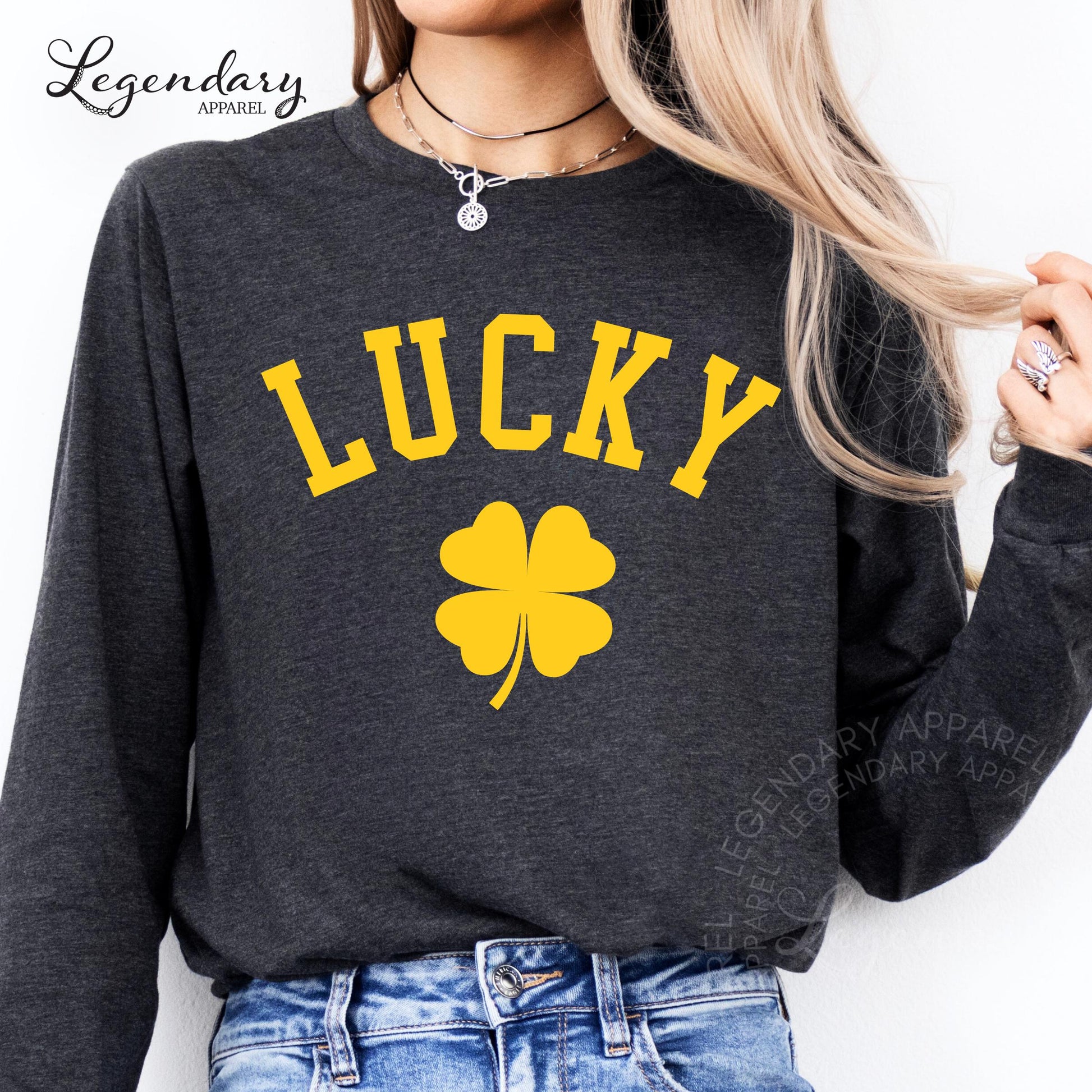 A woman wearing a gray sweatshirt with the word "Lucky" printed on it, along with a yellow clover symbol. She is also wearing a necklace and jeans.