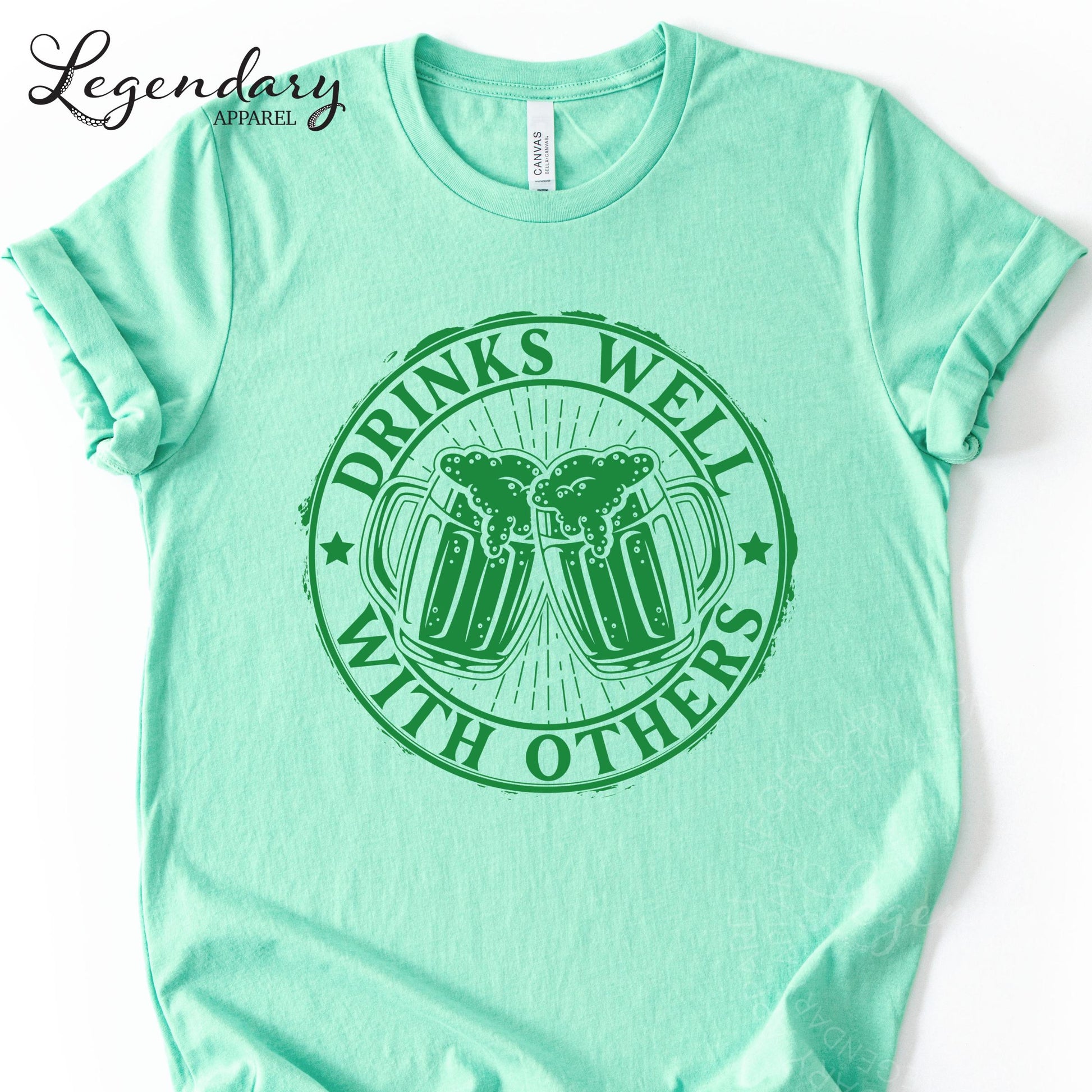 A mint green t-shirt with a graphic design featuring two beer mugs and the text "DRINKS WELL WITH OTHERS" printed on it.