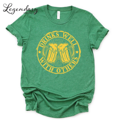 A green t-shirt with a graphic design that says "Drinks well with others" in yellow text.