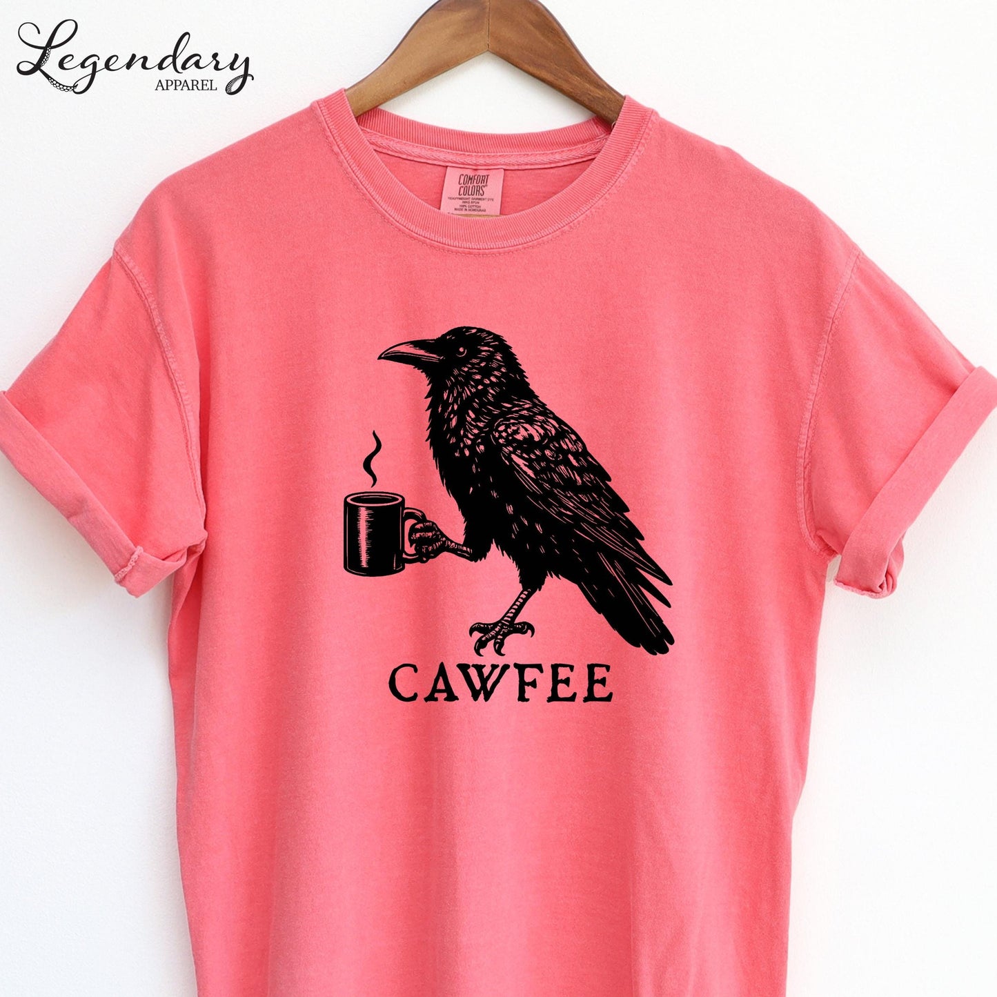 A pink t-shirt with a black illustration of a crow holding a coffee cup and the text "CAWFEE" below it.