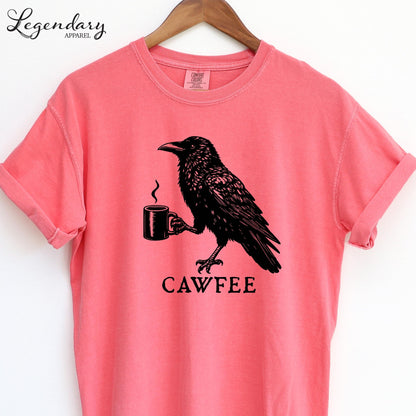 A pink t-shirt with a black illustration of a crow holding a coffee cup and the text "CAWFEE" below it.