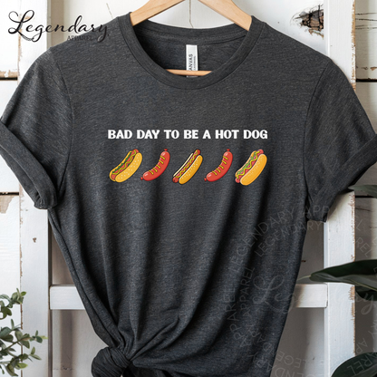 A gray t-shirt with a graphic design featuring four hot dogs in different colors and styles, along with the text "Bad day to be a hot dog".