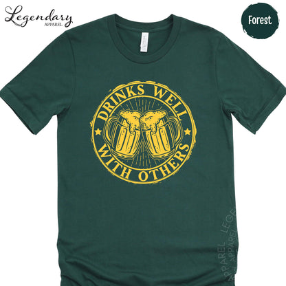 A green t-shirt with a graphic design featuring two beer mugs with a text message that reads "Drinks well with others".