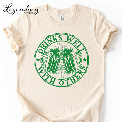 a white t-shirt with a green graphic design on it. The design consists of two beer mugs with a text that reads "DRINKS WELL WITH OTHERS".