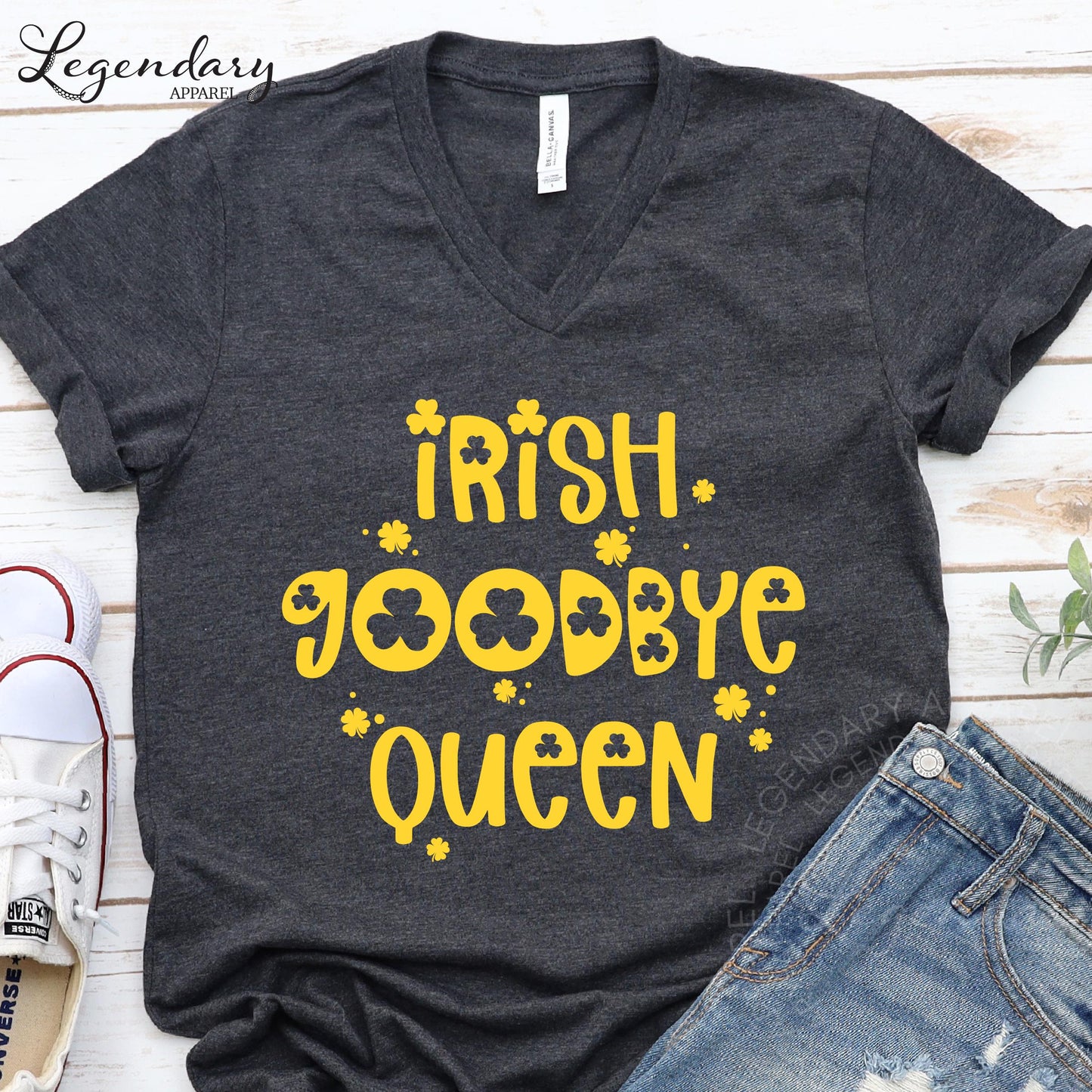 A gray t-shirt with yellow text and symbols, including the words "Irish. Goodbye Queen" and shamrocks, is displayed on a wooden surface alongside a pair of jeans and a pair of sneakers.