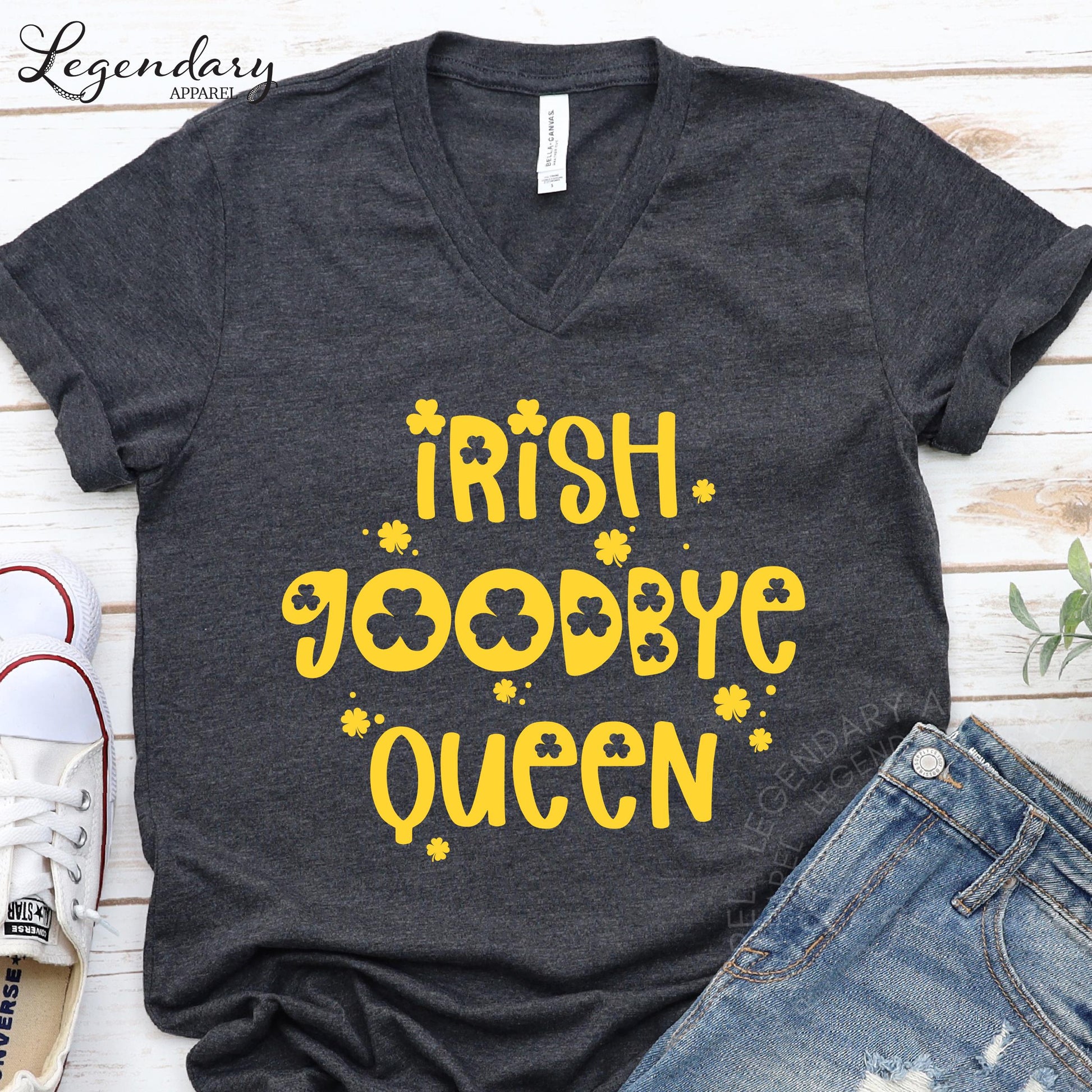A gray t-shirt with yellow text and symbols, including the words "Irish. Goodbye Queen" and shamrocks, is displayed on a wooden surface alongside a pair of jeans and a pair of sneakers.