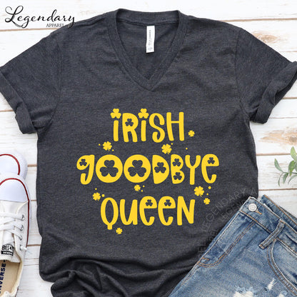 A gray t-shirt with yellow text and symbols, including the words "Irish. Goodbye Queen" and shamrocks, is displayed on a wooden surface alongside a pair of jeans and a pair of sneakers.