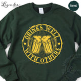 A green sweatshirt with a yellow graphic design on it. The design features two beer mugs with a text that says "DRINKS WELL WITH OTHERS".