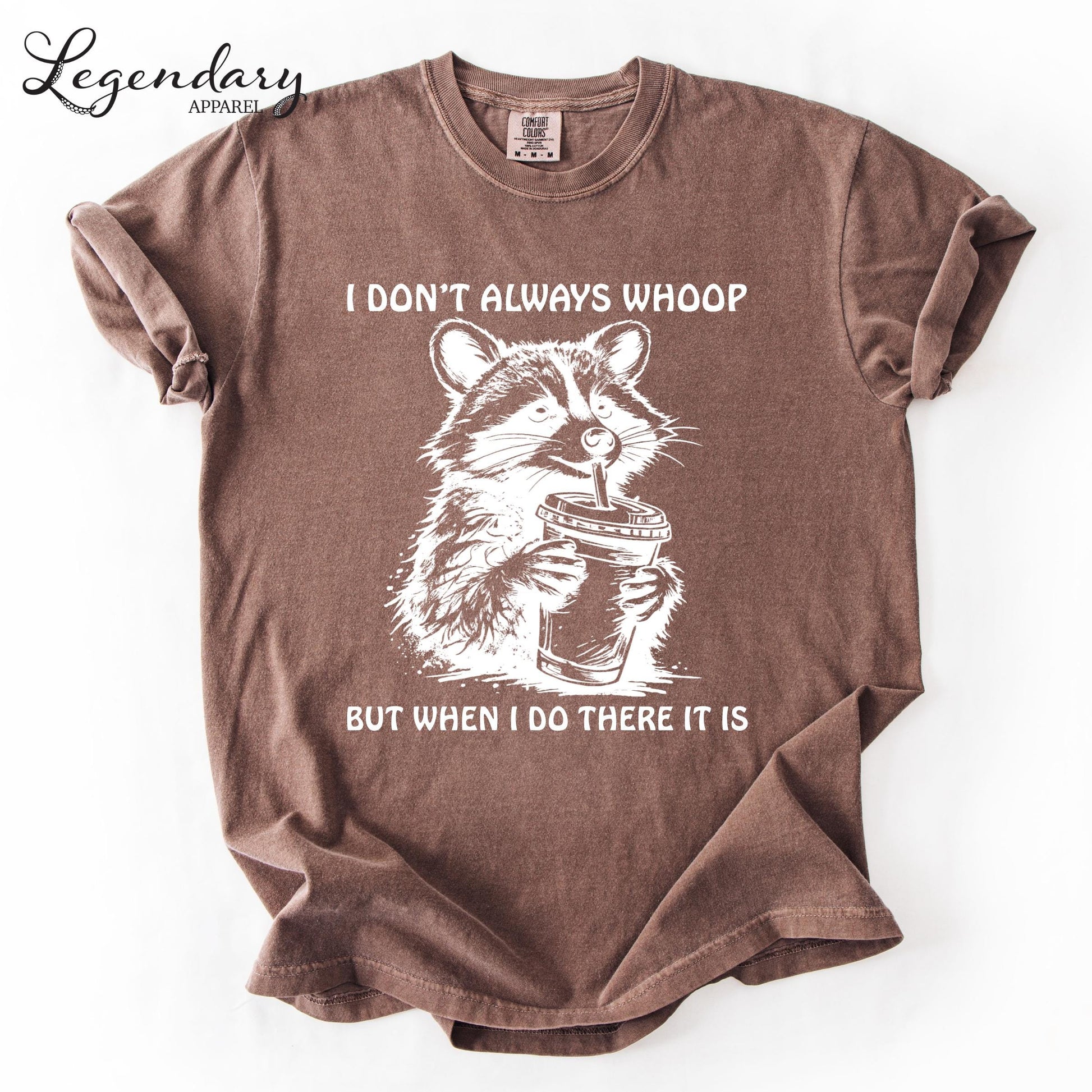 A brown t-shirt with a graphic design depicting a raccoon holding a cup, accompanied by the text "I DON'T ALWAYS WHOOP BUT WHEN I DO THERE IT IS".