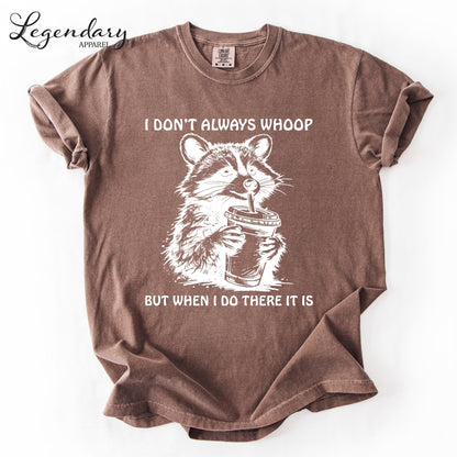 A brown t-shirt with a graphic design depicting a raccoon holding a cup, accompanied by the text "I DON'T ALWAYS WHOOP BUT WHEN I DO THERE IT IS".