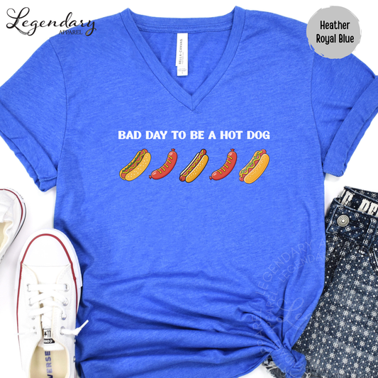 A blue t-shirt with the text "Bad day to be a hot dog" printed on it, along with illustrations of hot dogs. The t-shirt is placed on a white surface, and there are a pair of white sneakers and a pair of blue jeans visible in the image.