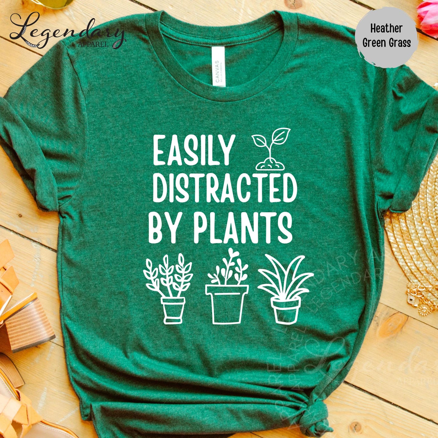 a green t-shirt with a humorous text printed on it, which reads "EASILY DISTRACTED BY PLANTS". The t-shirt is displayed on a wooden surface, and there are some potted plants visible in the background.