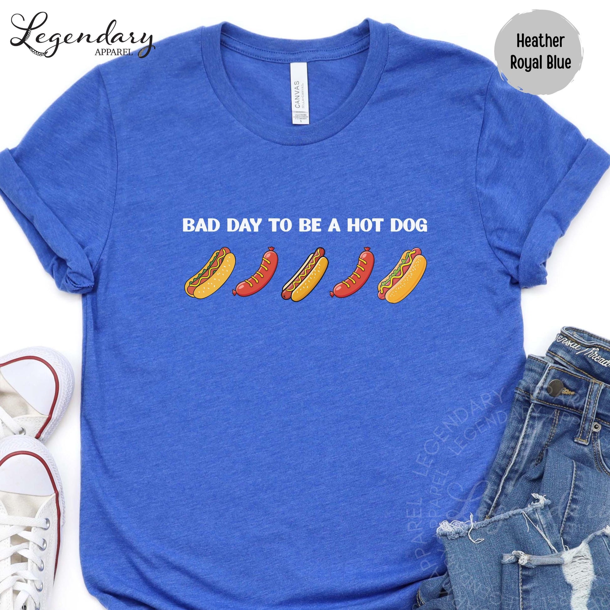 A blue t-shirt with the text "Bad day to be a hot dog" printed on it, along with illustrations of hot dogs. The t-shirt is placed next to a pair of jeans and sneakers.