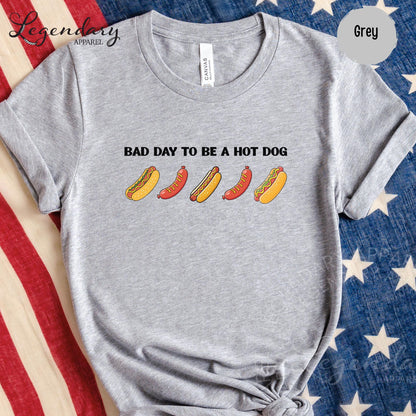 A grey t-shirt with a graphic design of five hot dogs in various colors and styles, along with the text "Bad day to be a hot dog".