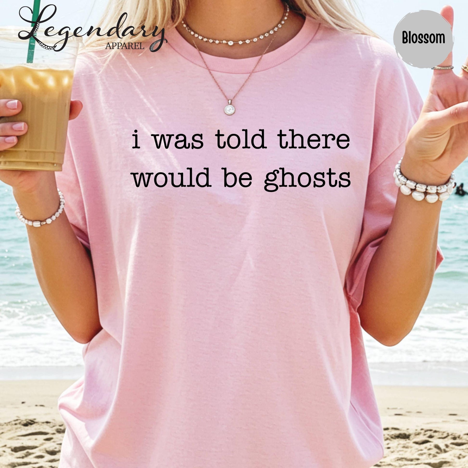 A person wearing a pink t-shirt with the text "I was told there would be ghosts" printed on it, standing on a beach with the ocean in the background.