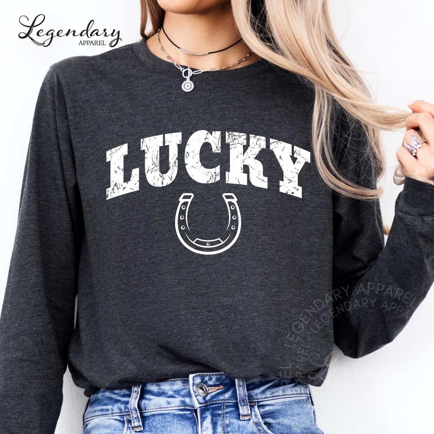 A woman wearing a grey long-sleeved shirt with the word "Lucky" printed on it, along with a horseshoe graphic. She is also wearing a necklace and jeans.