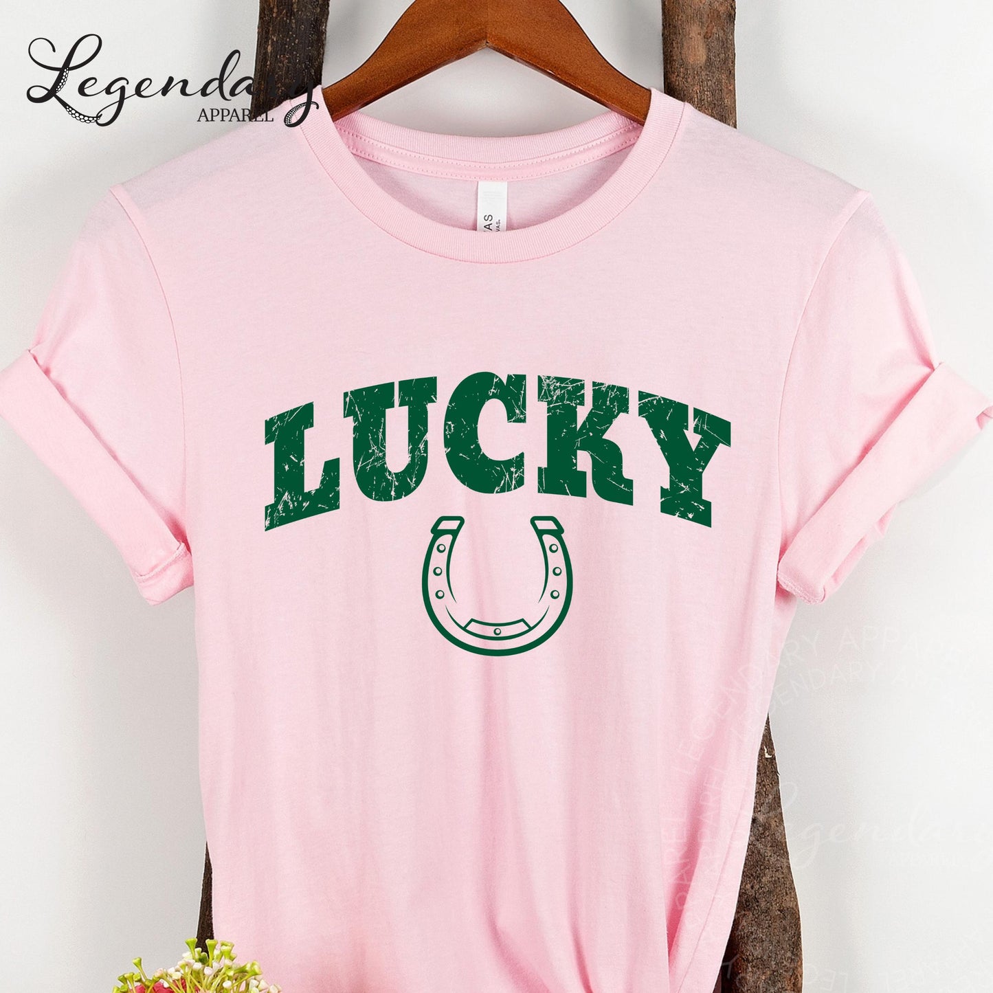 A pink t-shirt with the word "Lucky" printed in green letters, along with a horseshoe graphic, is hanging on a wooden hanger.