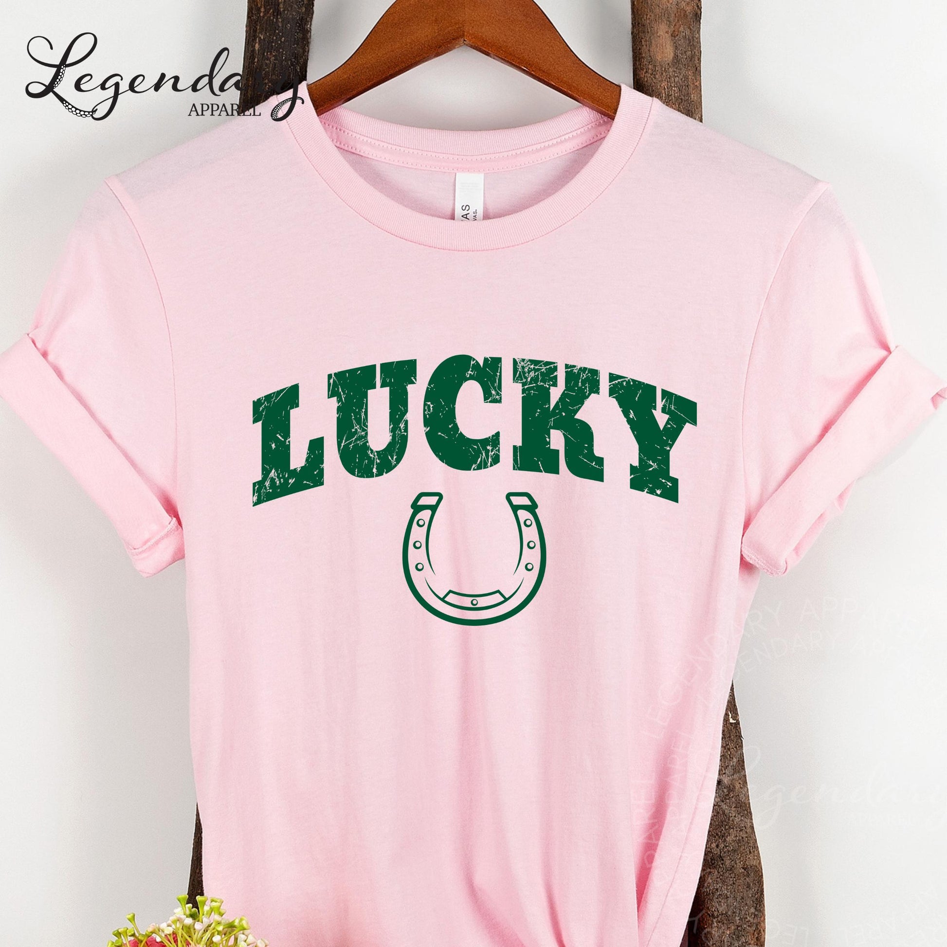 A pink t-shirt with the word "Lucky" printed in green letters, along with a horseshoe graphic, is hanging on a wooden hanger.