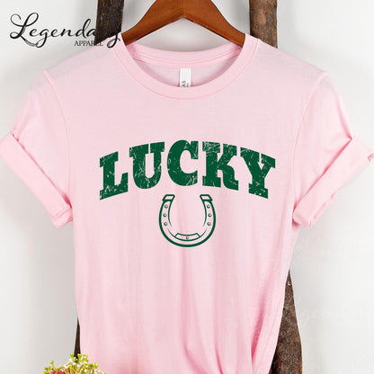 A pink t-shirt with the word "Lucky" printed in green letters, along with a horseshoe graphic, is hanging on a wooden hanger.
