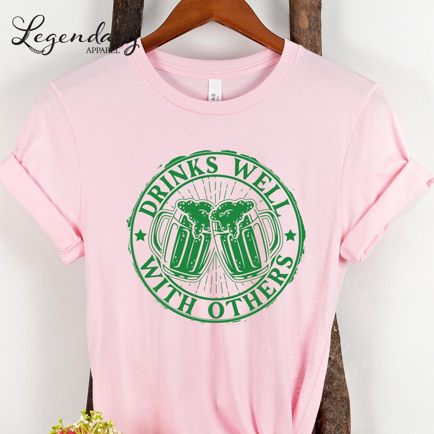 A pink t-shirt with a green graphic design that says "DRINKS WELCOME WITH OTHERS" on it.