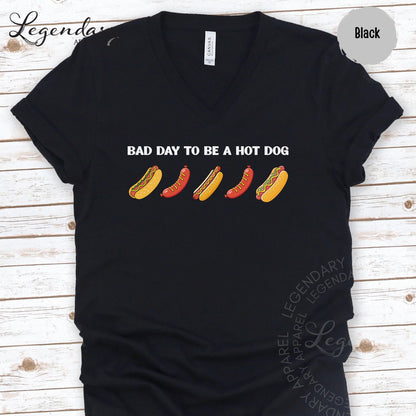 A black t-shirt with a graphic design featuring a line of hot dogs in various colors, accompanied by the text "Bad day to be a hot dog".