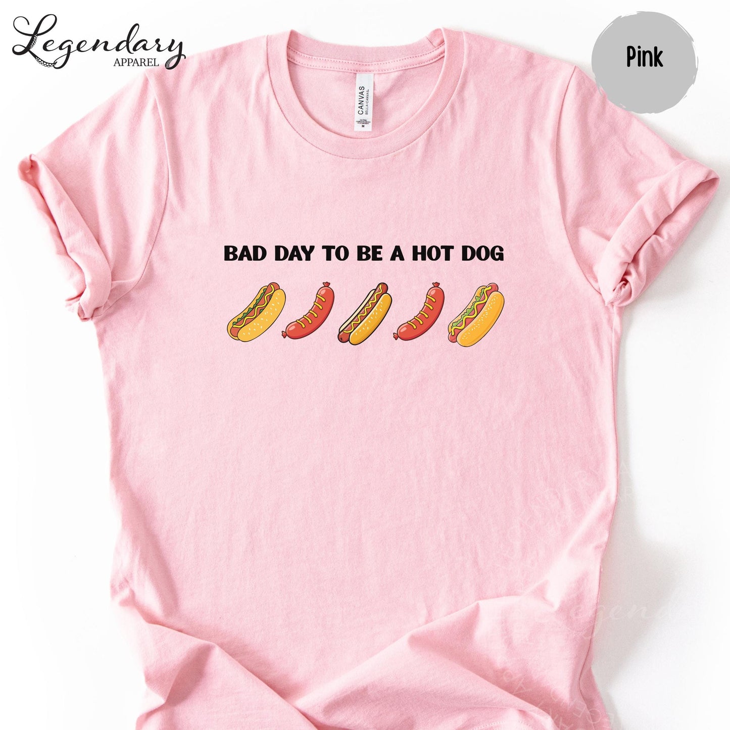 A pink t-shirt with a graphic design featuring five hot dogs in various colors and styles, along with the text "Bad day to be a hot dog".
