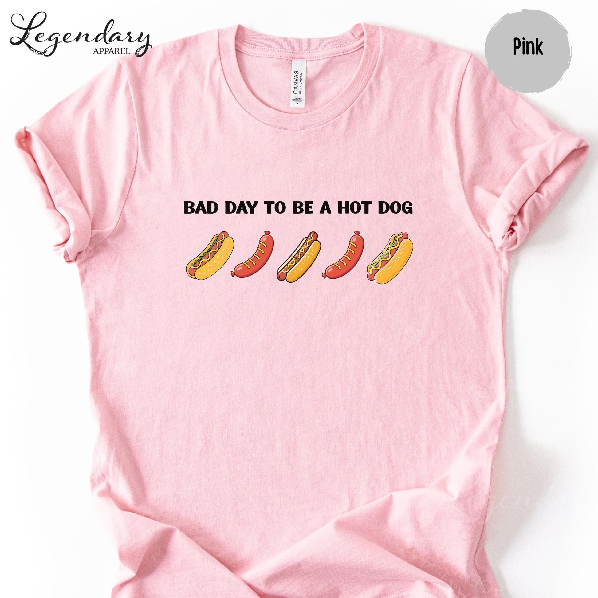 A pink t-shirt with a graphic design featuring five hot dogs in various colors and styles, along with the text "Bad day to be a hot dog".