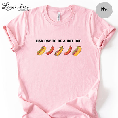 A pink t-shirt with a graphic design featuring five hot dogs in various colors and styles, along with the text "Bad day to be a hot dog".