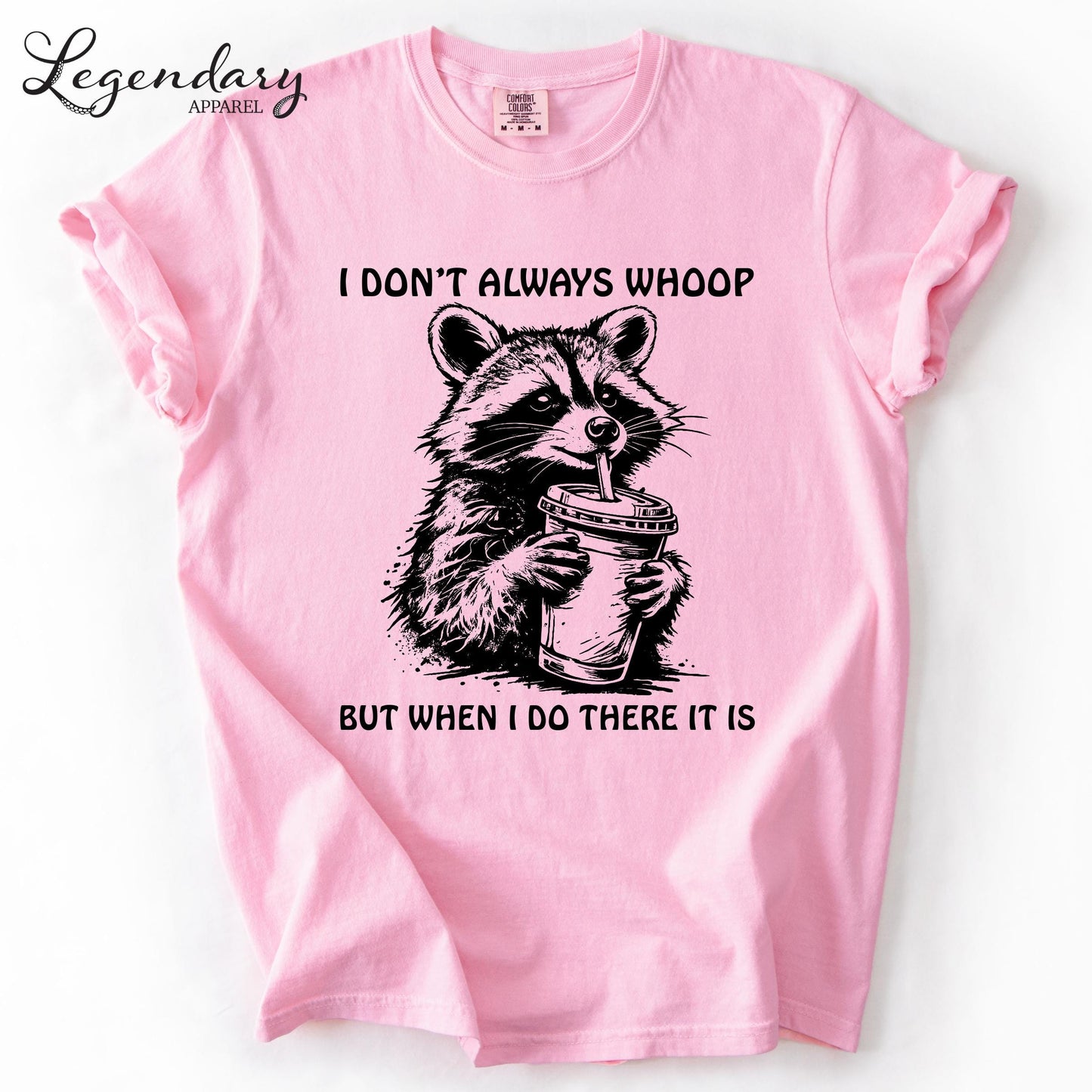 A pink t-shirt with a graphic design depicting a raccoon holding a cup, accompanied by the text "I DON'T ALWAYS WHOOP BUT WHEN I DO THERE IT IS".