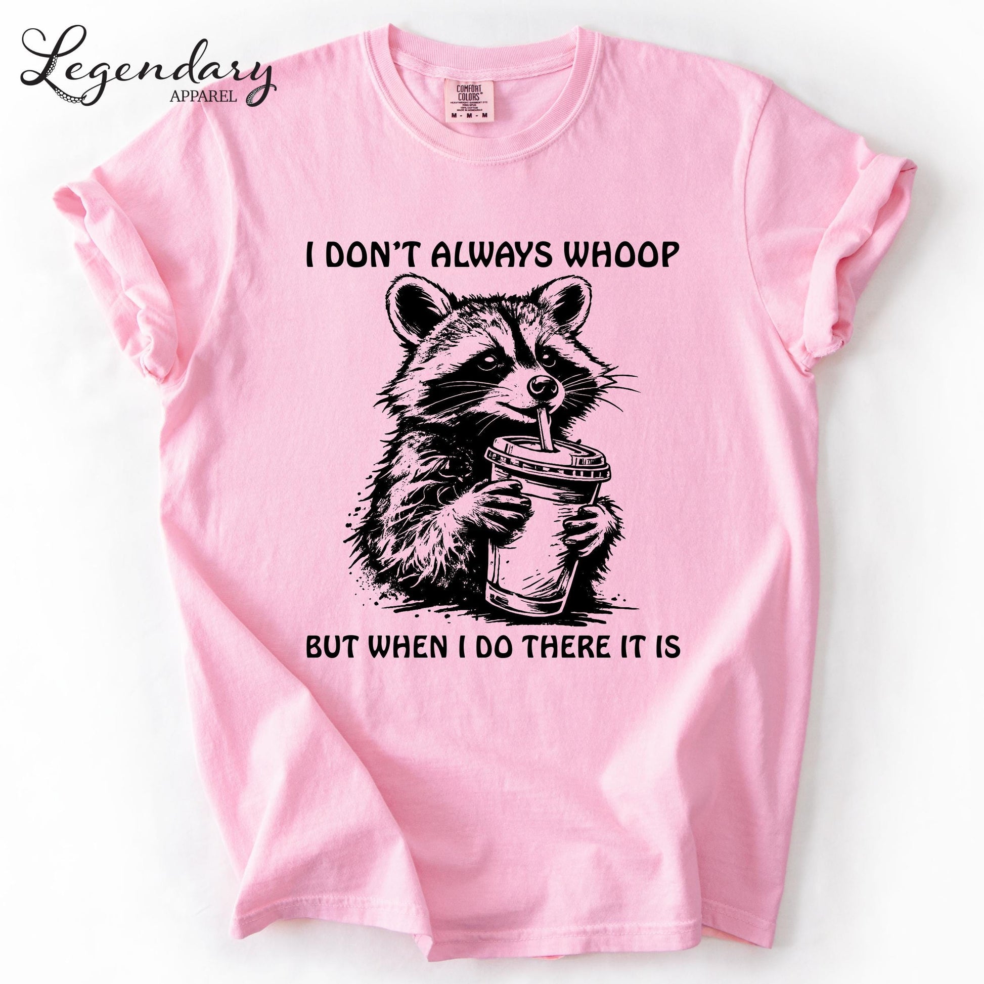 A pink t-shirt with a graphic design depicting a raccoon holding a cup, accompanied by the text "I DON'T ALWAYS WHOOP BUT WHEN I DO THERE IT IS".